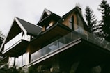 A modern house with a sharp, angular roofline features wooden paneling and large glass balconies. It is surrounded by tall trees, suggesting a forested location. The house has a contemporary design with natural materials, blending into the surroundings.