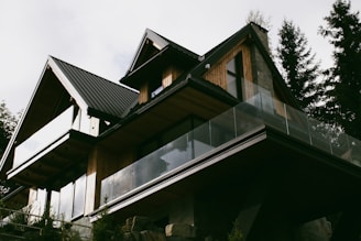 A modern house with a sharp, angular roofline features wooden paneling and large glass balconies. It is surrounded by tall trees, suggesting a forested location. The house has a contemporary design with natural materials, blending into the surroundings.