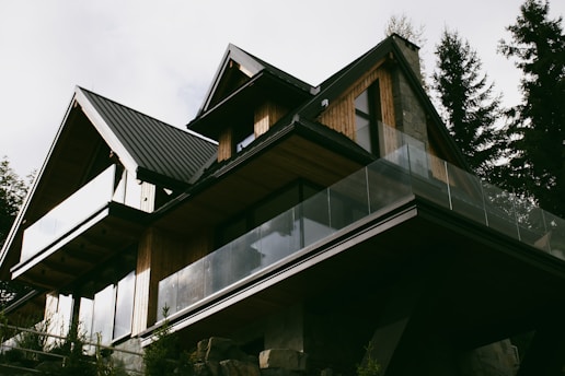 A modern house with a sharp, angular roofline features wooden paneling and large glass balconies. It is surrounded by tall trees, suggesting a forested location. The house has a contemporary design with natural materials, blending into the surroundings.