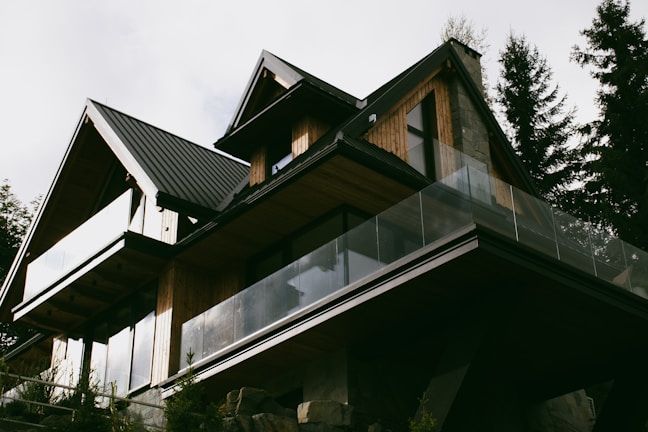 A modern house with a sharp, angular roofline features wooden paneling and large glass balconies. It is surrounded by tall trees, suggesting a forested location. The house has a contemporary design with natural materials, blending into the surroundings.