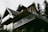 A modern house with a sharp, angular roofline features wooden paneling and large glass balconies. It is surrounded by tall trees, suggesting a forested location. The house has a contemporary design with natural materials, blending into the surroundings.