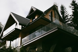 A modern house with a sharp, angular roofline features wooden paneling and large glass balconies. It is surrounded by tall trees, suggesting a forested location. The house has a contemporary design with natural materials, blending into the surroundings.