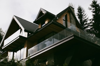 A modern house with a sharp, angular roofline features wooden paneling and large glass balconies. It is surrounded by tall trees, suggesting a forested location. The house has a contemporary design with natural materials, blending into the surroundings.