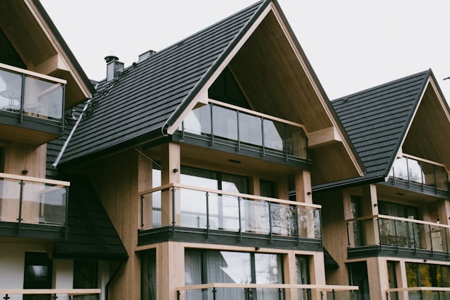 A series of modern wooden houses with large glass windows and triangular, black shingled roofs. The architecture features balconies with glass railings, creating a sleek and contemporary appearance.
