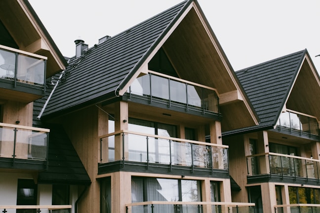 A series of modern wooden houses with large glass windows and triangular, black shingled roofs. The architecture features balconies with glass railings, creating a sleek and contemporary appearance.