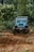A rugged off-road vehicle navigates through a muddy and uneven forest trail, surrounded by dense green foliage and trees on both sides.