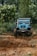 A rugged off-road vehicle navigates through a muddy and uneven forest trail, surrounded by dense green foliage and trees on both sides.