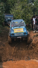 A blue off-road vehicle is attempting to climb a steep, muddy incline in a forested area. The vehicle is clearly engaged in an off-road adventure, with large muddy tires and a determined approach. Another vehicle is parked behind, while a group of individuals stand nearby, observing the event.