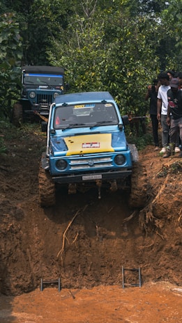 A blue off-road vehicle is attempting to climb a steep, muddy incline in a forested area. The vehicle is clearly engaged in an off-road adventure, with large muddy tires and a determined approach. Another vehicle is parked behind, while a group of individuals stand nearby, observing the event.