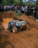 A blue off-road vehicle is being towed out of a muddy pit while a large crowd of people stand around watching. The scene is surrounded by dirt and some greenery in the background.