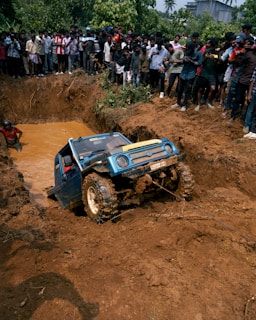 A blue off-road vehicle is being towed out of a muddy pit while a large crowd of people stand around watching. The scene is surrounded by dirt and some greenery in the background.
