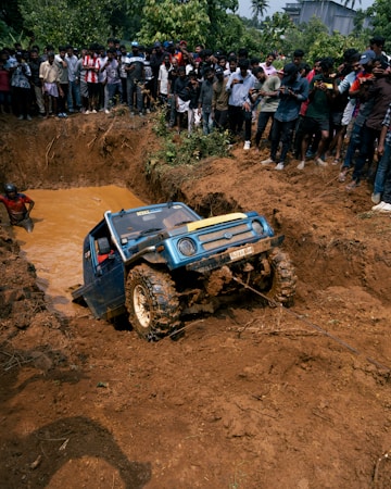 A blue off-road vehicle is being towed out of a muddy pit while a large crowd of people stand around watching. The scene is surrounded by dirt and some greenery in the background.