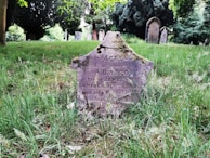A weathered and moss-covered gravestone stands in a cemetery, surrounded by green grass and tall trees. The stone has faded inscriptions and shows signs of age, with other gravestones visible in the background, suggesting a tranquil outdoor setting.