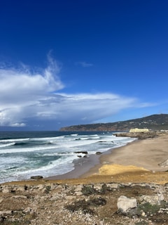 A picturesque beach with gentle waves rolling onto the sandy shore under a vibrant blue sky. The coastline stretches towards a distant rocky headland. A few clouds hover in the sky, adding depth to the scene. On the right, there is a building near the edge of the beach, with hills in the background.