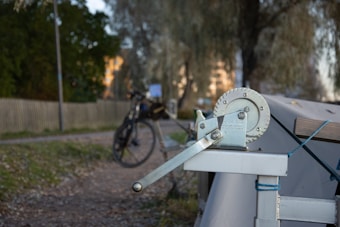 A close-up of a mechanical hand winch mounted on a structure with a bicycle and a park setting in the background. The background also features trees, a fence, and some buildings.
