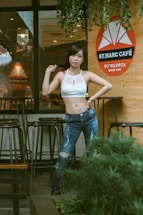 A young woman stands confidently in front of a cafe, posing with one hand on her hip and the other holding her hair. She wears a white crop top and blue distressed jeans. The cafe features a wooden wall with a prominent orange and white logo that reads 'ST. MARC CAFE'. There are tall bar stools and potted greenery nearby, adding to the inviting outdoor atmosphere.