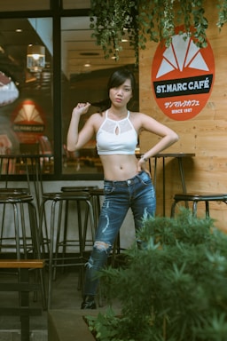 A young woman stands confidently in front of a cafe, posing with one hand on her hip and the other holding her hair. She wears a white crop top and blue distressed jeans. The cafe features a wooden wall with a prominent orange and white logo that reads 'ST. MARC CAFE'. There are tall bar stools and potted greenery nearby, adding to the inviting outdoor atmosphere.