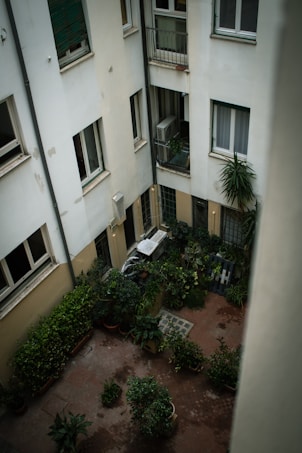 A small courtyard surrounded by several building walls, featuring numerous potted plants and greenery arranged neatly. The space includes a mix of windows and small balconies, with an air conditioning unit visible. The overall atmosphere appears calm and cozy.