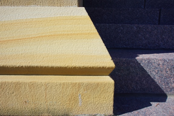 A section of a stairway with textured surfaces, featuring a contrast between rough yellow and gray stone textures. The area is bisected by a sharp shadow, creating a geometric pattern.