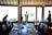 A dimly lit room with a large window showcasing a silhouette of a man holding skis, along with the number '007' on the glass. Inside, there are several tables set with glassware and placemats, with three people present—one seated, another facing the window, and a woman standing to the side.