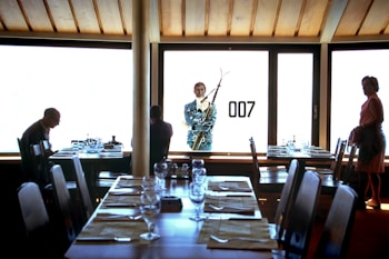 A dimly lit room with a large window showcasing a silhouette of a man holding skis, along with the number '007' on the glass. Inside, there are several tables set with glassware and placemats, with three people present—one seated, another facing the window, and a woman standing to the side.