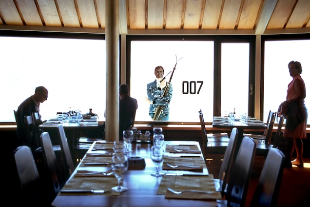 A dimly lit room with a large window showcasing a silhouette of a man holding skis, along with the number '007' on the glass. Inside, there are several tables set with glassware and placemats, with three people present—one seated, another facing the window, and a woman standing to the side.