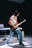 A person is energetically playing a guitar in a dimly lit room. They are wearing a colorful shirt and jeans, and their posture is expressive. In the background, there's a table with a laptop covered in stickers, and a chair. The spotlight highlights the musician, creating a dramatic effect.