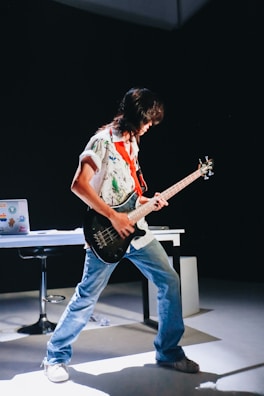 A person is energetically playing a guitar in a dimly lit room. They are wearing a colorful shirt and jeans, and their posture is expressive. In the background, there's a table with a laptop covered in stickers, and a chair. The spotlight highlights the musician, creating a dramatic effect.
