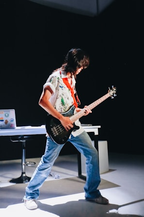 A person is energetically playing a guitar in a dimly lit room. They are wearing a colorful shirt and jeans, and their posture is expressive. In the background, there's a table with a laptop covered in stickers, and a chair. The spotlight highlights the musician, creating a dramatic effect.