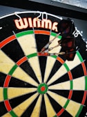 A dartboard with several darts sticking into the bullseye, surrounded by a pattern of red, green, and beige sections. The brand name is visible at the top.