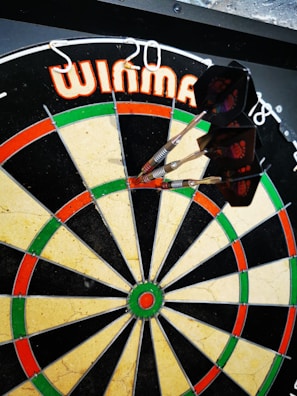 A dartboard with several darts sticking into the bullseye, surrounded by a pattern of red, green, and beige sections. The brand name is visible at the top.