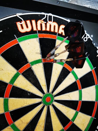 A dartboard with several darts sticking into the bullseye, surrounded by a pattern of red, green, and beige sections. The brand name is visible at the top.