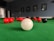 A close-up view of a billiard table with a white cue ball prominently positioned in the foreground. Several red balls and a single black ball are scattered across the green felt surface. The background is out of focus, featuring framed pictures on the wall, contributing to the ambiance of a billiards room.