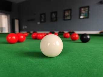 A close-up view of a billiard table with a white cue ball prominently positioned in the foreground. Several red balls and a single black ball are scattered across the green felt surface. The background is out of focus, featuring framed pictures on the wall, contributing to the ambiance of a billiards room.