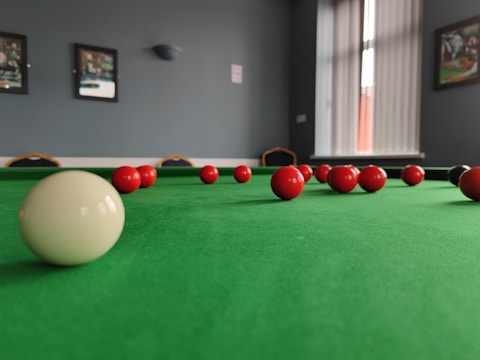 A snooker table with a green felt surface is seen in a room with framed pictures on the walls. Several red balls and one white ball are positioned on the table, capturing the depth and perspective towards a window with vertical blinds.