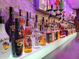 A well-organized bar shelf displaying a variety of liquor bottles, including brands like Bailey’s, Captain Morgan, and Jack Daniel’s. The bottles are arranged on a glowing white surface with a lit brick wall background. Some bottles are mounted on dispensers above.