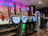 A bar setting featuring various beer taps prominently displayed in the foreground. The taps bear logos for different beer brands including Worthington's, Carling, Caffrey's, and Foster's. The background includes a variety of liquor bottles on shelves, illuminated by soft blue and purple lighting, adding a modern and inviting atmosphere.