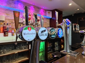 A bar setting featuring various beer taps prominently displayed in the foreground. The taps bear logos for different beer brands including Worthington's, Carling, Caffrey's, and Foster's. The background includes a variety of liquor bottles on shelves, illuminated by soft blue and purple lighting, adding a modern and inviting atmosphere.