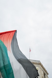 A flag with red, black, white, and green colors waves in the foreground. In the background, a building is visible with an American flag on top against a cloudy sky.