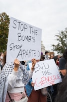 A group of people are holding protest signs with messages such as 'Stop the Bombs, Stop Apartheid' and 'End the Genocide' in an outdoor setting. The crowd appears to be engaged in a demonstration or rally. Some individuals are wearing patterned scarves, and there are trees and part of a large building visible in the background.