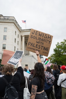 A crowd of people gathered outside a government building, some holding signs expressing solidarity with Palestine and calling for a ceasefire. The American flag is visible on top of the building, and trees are in the background.