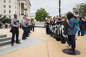 A group of police officers stands in a line facing a crowd of protestors holding signs and banners in front of a large, columned building. The protestors hold a black banner with bold white letters. The scene appears to be tense, with clear separation between the officers and protestors.