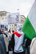 A group of people gathers for a protest, holding signs related to the conflict in Palestine. One poster depicts an illustration involving political themes, and the central figure wears a clothing item displaying the colors of the Palestinian flag. The scene unfolds near a historical building with an American flag visible in the background.