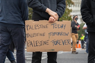 A person stands holding a cardboard sign with the words 'Palestine Today, Palestine Tomorrow, Palestine Forever' written on it. The scene appears to be part of a protest or demonstration. There are other people in the background, some wearing masks, and part of a cityscape is visible.