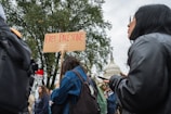 A group of people participating in a protest. One individual is holding a cardboard sign with the text 'FREE PALESTINE'. They are standing in front of a large tree, and in the background, there is a recognizable dome-shaped government building.