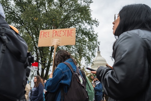 A group of people participating in a protest. One individual is holding a cardboard sign with the text 'FREE PALESTINE'. They are standing in front of a large tree, and in the background, there is a recognizable dome-shaped government building.