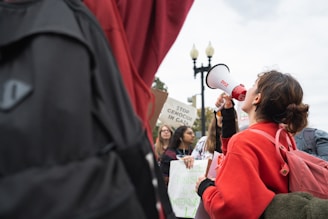 A person wearing a red jacket is holding a megaphone, surrounded by a group of people at a protest. Several signs are visible, with messages regarding genocide and international law. The setting appears to be outdoors with overcast skies and a lamp post in the background.