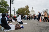 A group of people gather in front of a large, iconic white building with a dome, holding signs and engaging in activities that suggest a protest or demonstration. Some individuals are seated on blankets while others stand holding placards. The atmosphere is busy, with trees and streetlights visible in the background.