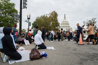 A group of people gather in front of a large, iconic white building with a dome, holding signs and engaging in activities that suggest a protest or demonstration. Some individuals are seated on blankets while others stand holding placards. The atmosphere is busy, with trees and streetlights visible in the background.