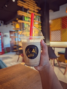 A hand holds a chilled drink in a plastic cup with a red straw, featuring a logo on the cup. The background shows a modern cafe with stylish furniture and decorative elements like wooden slats and plants.